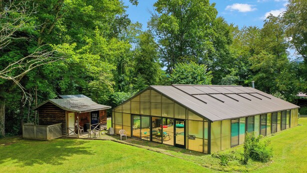 Een modern glazen huis met een terras en een houten blokhut in een weelderige, groene omgeving.
