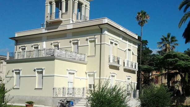 A two-story building with a central tower, balconies, and a small garden area with a palm tree. - Beachfront apartment with garden (Grottammare)