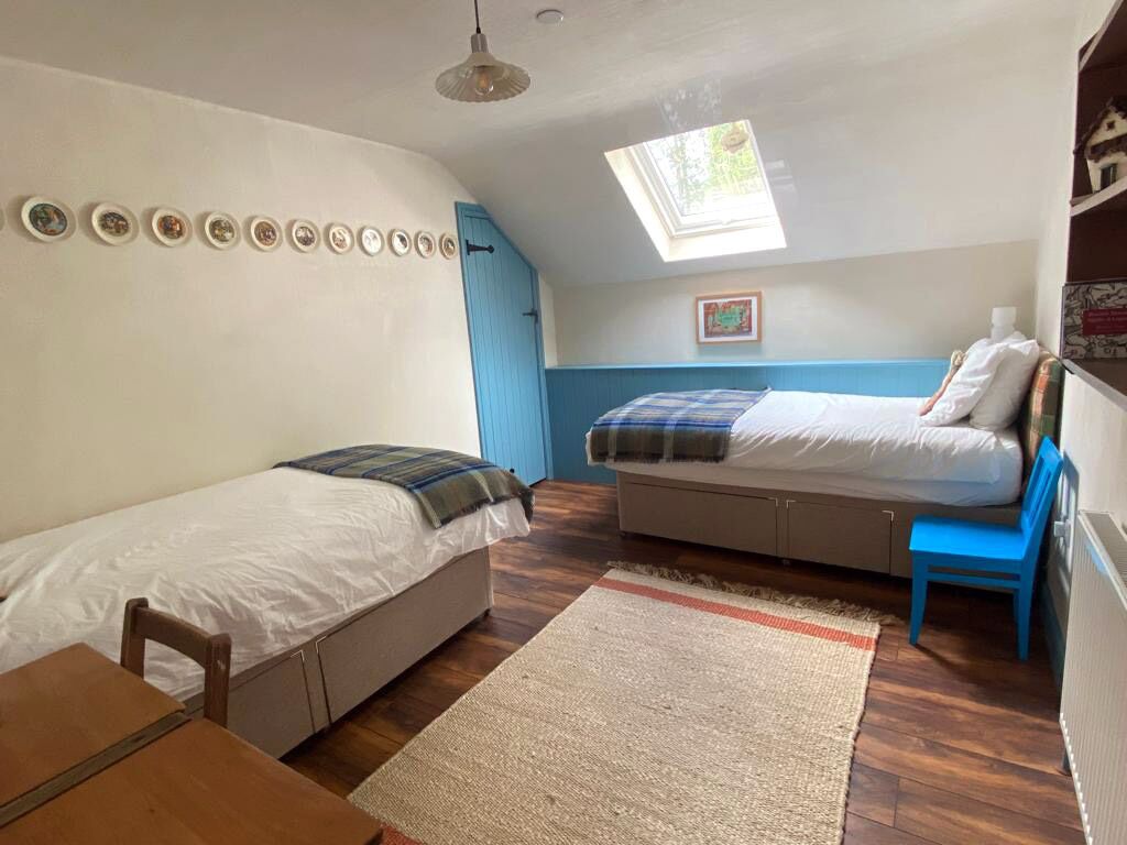 A small, cozy room with two single beds, a wooden table, a blue chair, a skylight, and a wall-mounted shelf with decorative items.