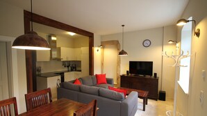 En moderne stue med en grå sofa, et træspisebord og stole. Et fjernsyn er monteret på væggen, og der er et køkken i baggrunden.