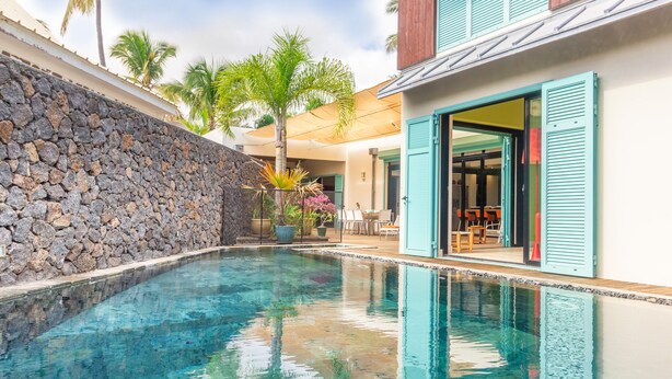 A swimming pool with a stone wall, a wooden deck, and a building with teal shutters. - La Musarde, magnificent villa with swimming pool close to the beach of Boucan Canot (Boucan Canot)