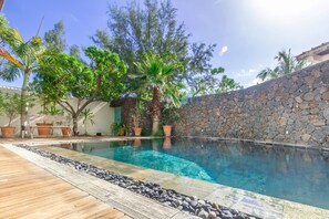 A swimming pool with a stone wall and wooden deck, surrounded by potted plants and trees.