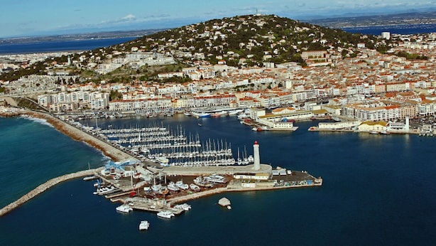 En vy från ovan över en kuststad med en småbåtshamn, byggnader och en kulle i bakgrunden.