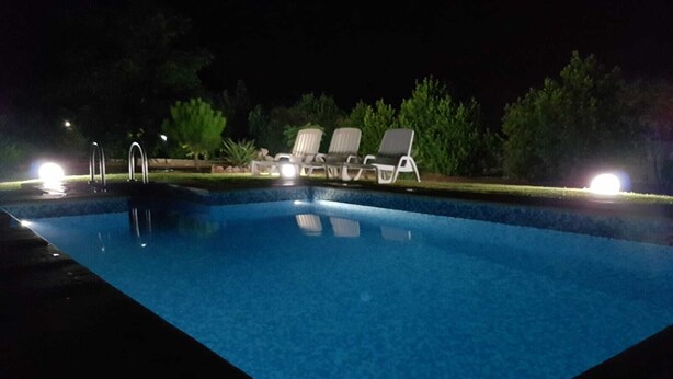 Una piscina por la noche con tres sillas de descanso y un pequeño jardín.
