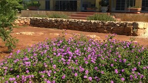 Un edificio amarillo con tejado de tejas, muros de piedra y un jardín con flores moradas.