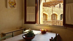 A dining area with a table set for two, a window with an open shutter, and a view of a stone building with arches.