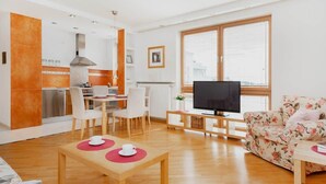 Una sala moderna con suelo de madera, un sofá floral, una zona de comedor con sillas blancas, una cocina con azulejos naranjas y un ventanal con persianas.