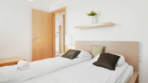 Habitación de hotel con dos camas, cabecero de madera, una mesita de noche y una pequeña estantería en la pared con un jarrón, y una puerta que da a otra habitación.