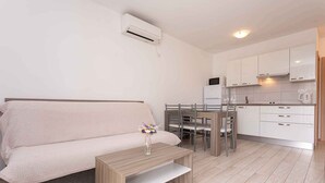 Un espacio habitable moderno y minimalista con sofá, una pequeña mesa de madera, comedor con sillas y cocina con armarios y electrodomésticos blancos.