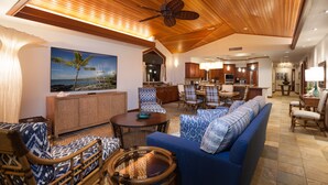 A spacious living area with a large sofa, wicker chairs, and a dining table. A painting of a beach scene is on the wall, and the room has wooden ceilings and a ceiling fan. - Private Villa, Golf Course Front with a Private Pool and Spa! (Kamuela)