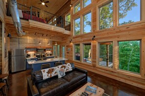 A spacious cabin interior with a kitchen, dining area, and a large window overlooking the forest.