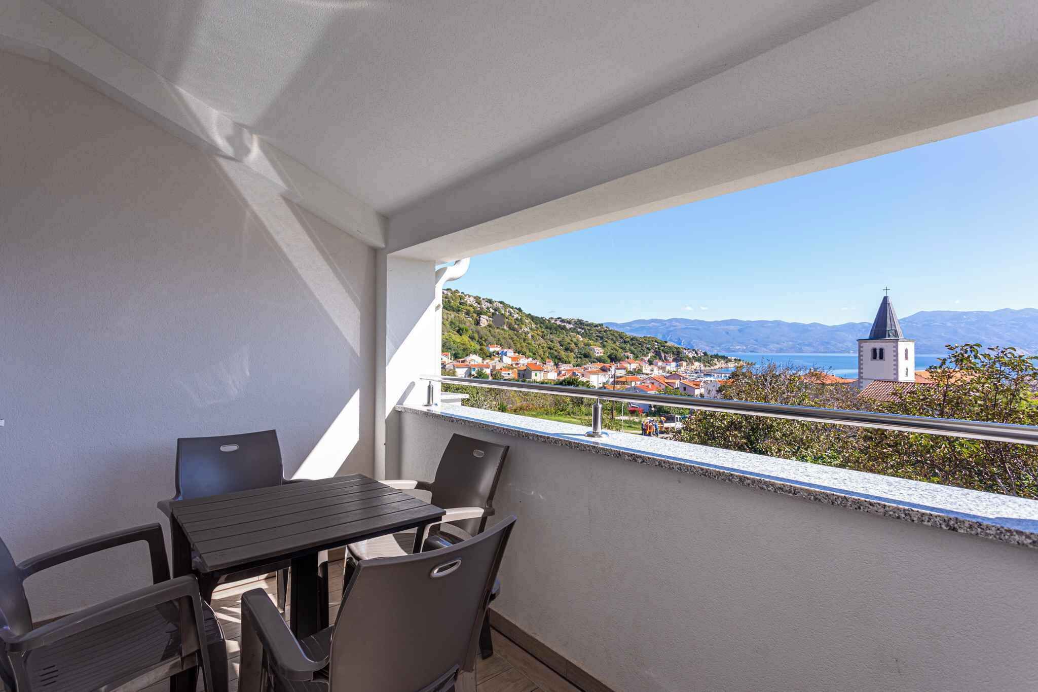 A balcony with a table and chairs, overlooking a town with a church and mountains.