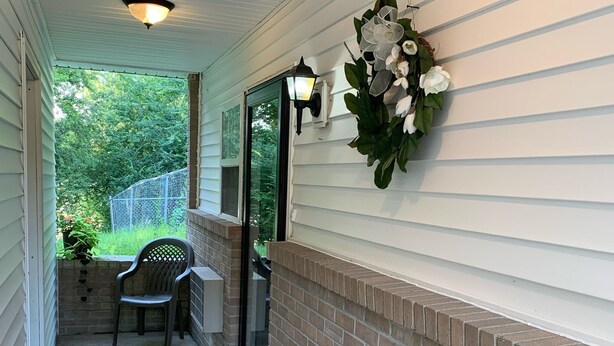 A covered porch with a brick wall, a chair, a potted plant, and a decorative wall hanging.