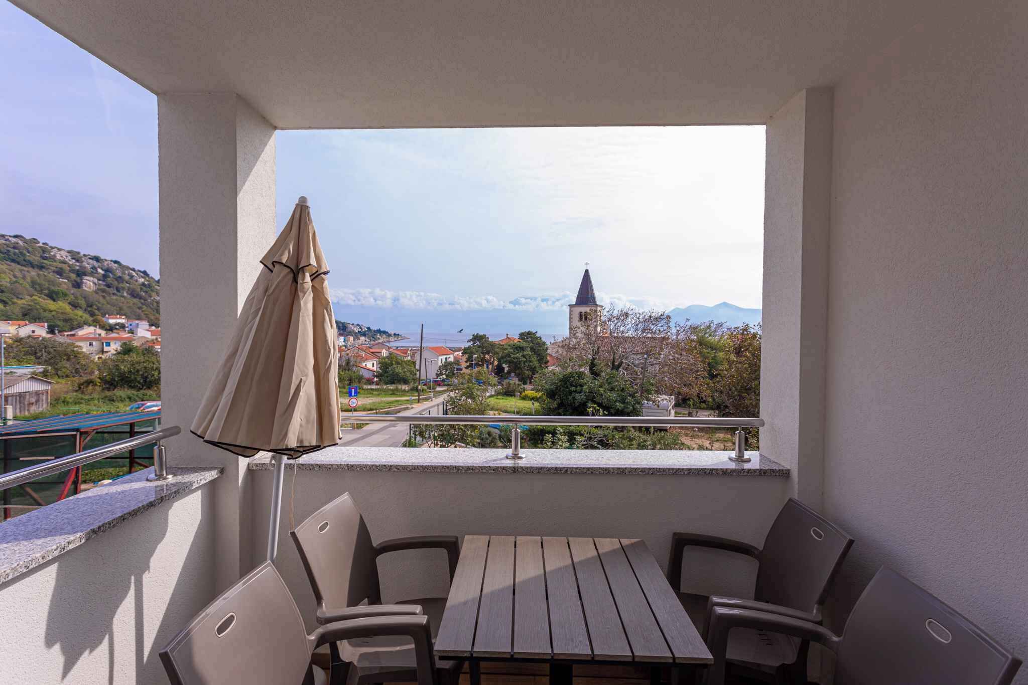 Un balcon avec une table et des chaises, un parasol beige, et une vue sur une ville avec une église et des montagnes à l’horizon.