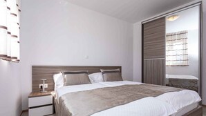 Un dormitorio moderno con cabecera de madera, una cama grande con manta color beige, una mesita de noche con lámpara y un armario con puerta corredera.