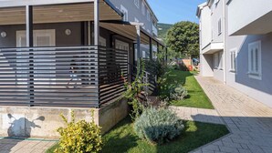 Un edificio residencial con un área exterior techada, un sendero pavimentado y un pequeño jardín con arbustos y césped.