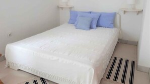 A neatly made bed with a white duvet and blue pillows, a framed painting above it, and a striped rug on the floor.
