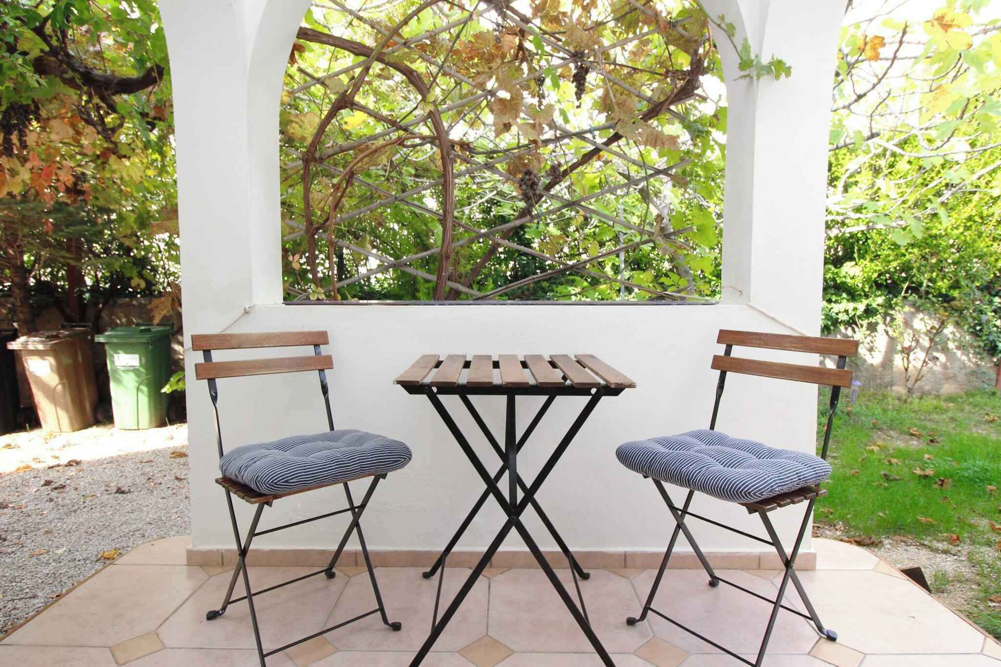 A small outdoor patio with a white wall, a wooden table, two foldable chairs, and a view of trees through a window.
