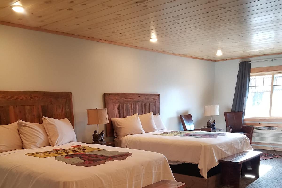 A hotel room with two beds, wooden headboards, a bench, a small table, and a window with curtains.