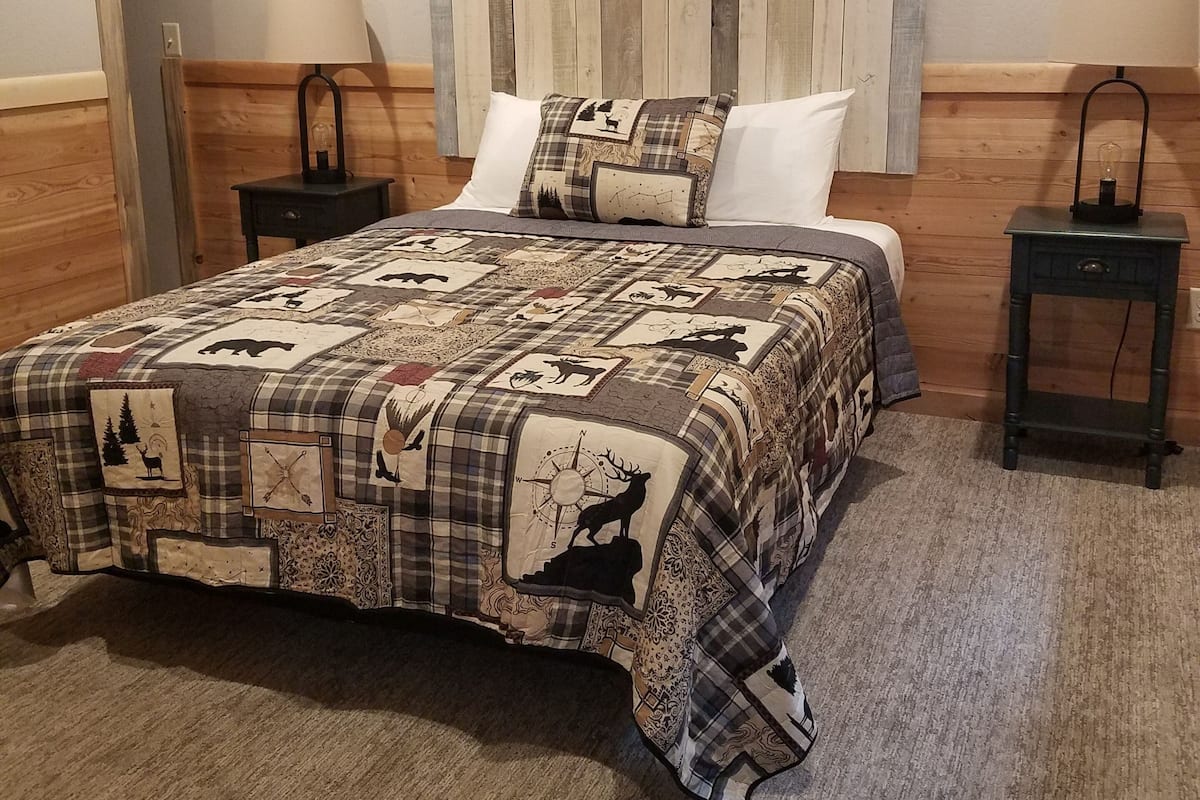 A bedroom with a wooden headboard, a quilted bedspread with animal prints, two bedside tables with lamps, and a carpeted floor.