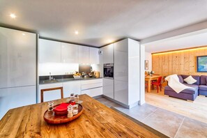 Une cuisine moderne avec des meubles blancs, une table à manger en bois sur laquelle est posé un plateau d’assaisonnements, et un coin salon chaleureux avec un canapé et un mur lambrissé.