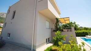 Ein modernes, zweistöckiges Gebäude mit Flachdach, einem Balkon mit gelbem Markisenstoff und einem Schwimmbad im Hintergrund.