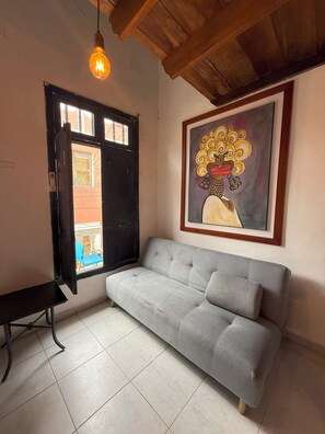 A room with a grey sofa, a black-framed window, a hanging light bulb, and a framed artwork on the wall.