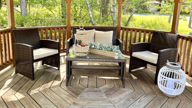A wooden gazebo with wicker furniture and a glass table, surrounded by a wooden deck and lush greenery.