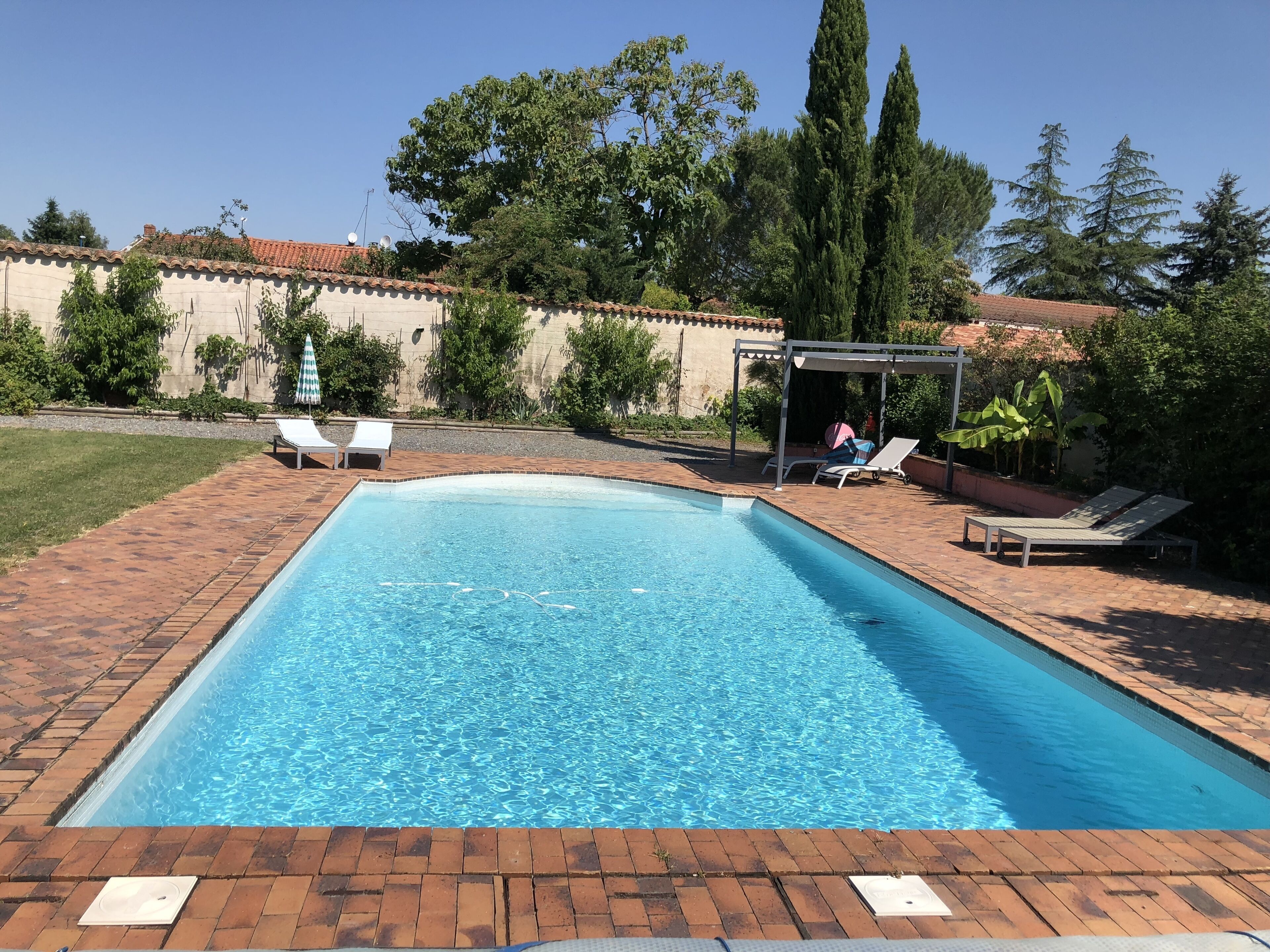 Une piscine rectangulaire dont les bords sont recouverts de carreaux, entourée d’une terrasse en briques. On y trouve des chaises longues et un espace de détente couvert avec une table.