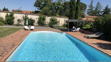 A rectangular swimming pool with a tiled edge, surrounded by a brick patio. There are lounge chairs and a covered seating area with a table.