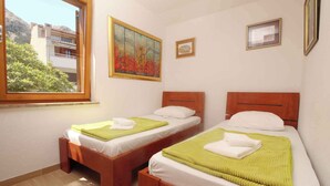 A room with two wooden beds, each with a green blanket and white pillows. There is a painting on the wall and a window with a view of buildings and trees.