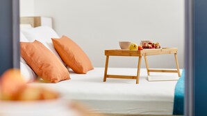 Ein ordentlich bezogenes Bett mit weißen Leinen und orangen Kissen, ein kleiner Holztisch mit Schüssel und Früchten sowie ein Blick auf einen Raum mit blauer Akzentwand.