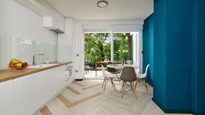Eine moderne Küche mit weißen Schränken, einer Holz Arbeitsplatte und einer Obstschale. Es gibt einen Essbereich mit Tisch und Stühlen sowie einen Blick ins Freie durch große Fenster.