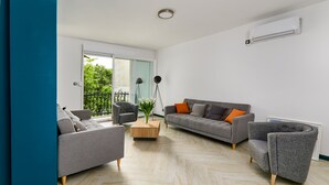 Ein modernes Wohnzimmer mit grauem Sofa, einem grauen Sessel und einem Holztisch. Ein großes Fenster bietet Blick ins Grüne.
