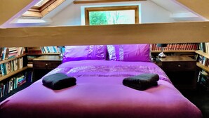 Un dormitorio con techo inclinado, tragaluz, cama con ropa de cama púrpura, dos almohadas y mesita de noche con lámpara.