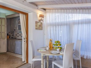 Un coin repas avec une table dressée pour deux personnes, une cuisine visible à travers une porte ouverte et une grande fenêtre ornée de rideaux légers.