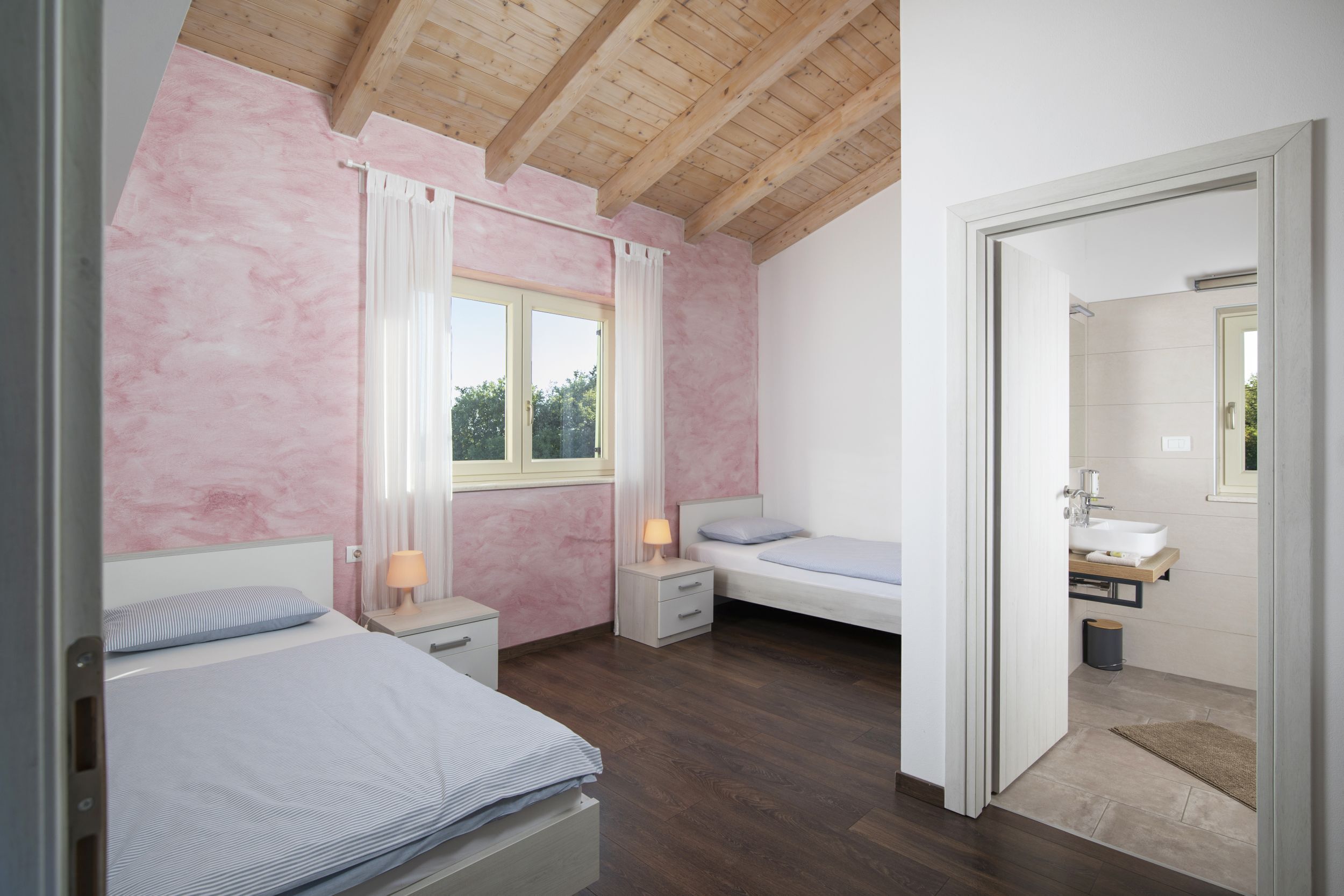Une chambre avec un plafond en bois, une fenêtre avec des rideaux, une table de chevet avec une lampe, un lit avec un oreiller et une vue sur une salle de bain.