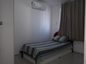 Una habitación pequeña con cama individual, colcha a rayas, unidad de aire acondicionado en la pared y ventana con cortinas.