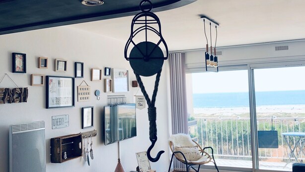 Una sala de estar moderna con un amplio ventanal que da a la playa, una lámpara colgante y un televisor montado en la pared.
