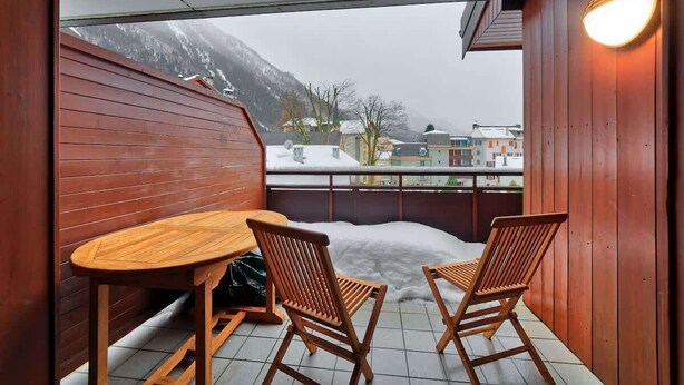 En balkong i trä med ett bord och två stolar, med utsikt över ett snötäckt berg och byggnader.