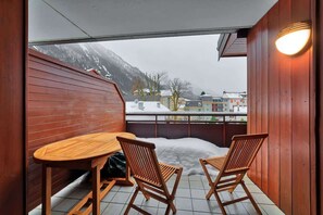 Ein Holzbalkon mit Tisch und zwei Stühlen, mit Blick auf einen verschneiten Berg und Gebäude.
