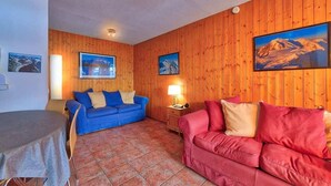 Ett vardagsrum med träpanel på väggarna, en blå soffa, en röd soffa och ett matbord med en duk på.