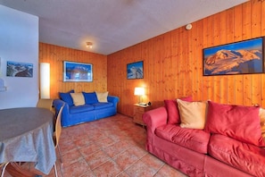 A living room with wooden walls, a blue sofa, a red sofa, and a dining table with a tablecloth.