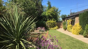 Um jardim bem cuidado com diversas plantas e flores, um banco de madeira e um edifício de tijolos ao fundo.
