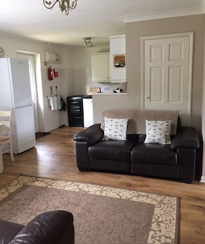 Een woonkamer met een donker leren bank, gekleurde kussens en een vloerkleed met patroon. Achtergrond toont een keuken met een koelkast, fornuis en keukenkastjes.