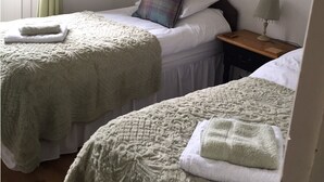 Quarto com duas camas, cada uma com um cobertor acolchoado e uma toalha dobrada. Há uma mesa de cabeceira com uma luminária, uma obra de arte na parede e uma janela com cortinas.