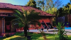 Una casa con techo rojo, un pequeño jardín con césped y una palmera, y un área de piscina con sillas de descanso.