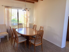 A dining area with a round wooden table surrounded by six wooden chairs with cushioned seats. A white sofa is positioned against the wall, and a window with sheer curtains allows natural light to enter.