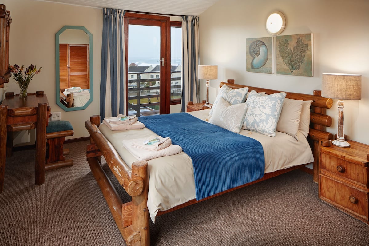 A hotel room with a wooden bed, a desk, a chair, a mirror, a window with curtains, and artwork on the wall.
