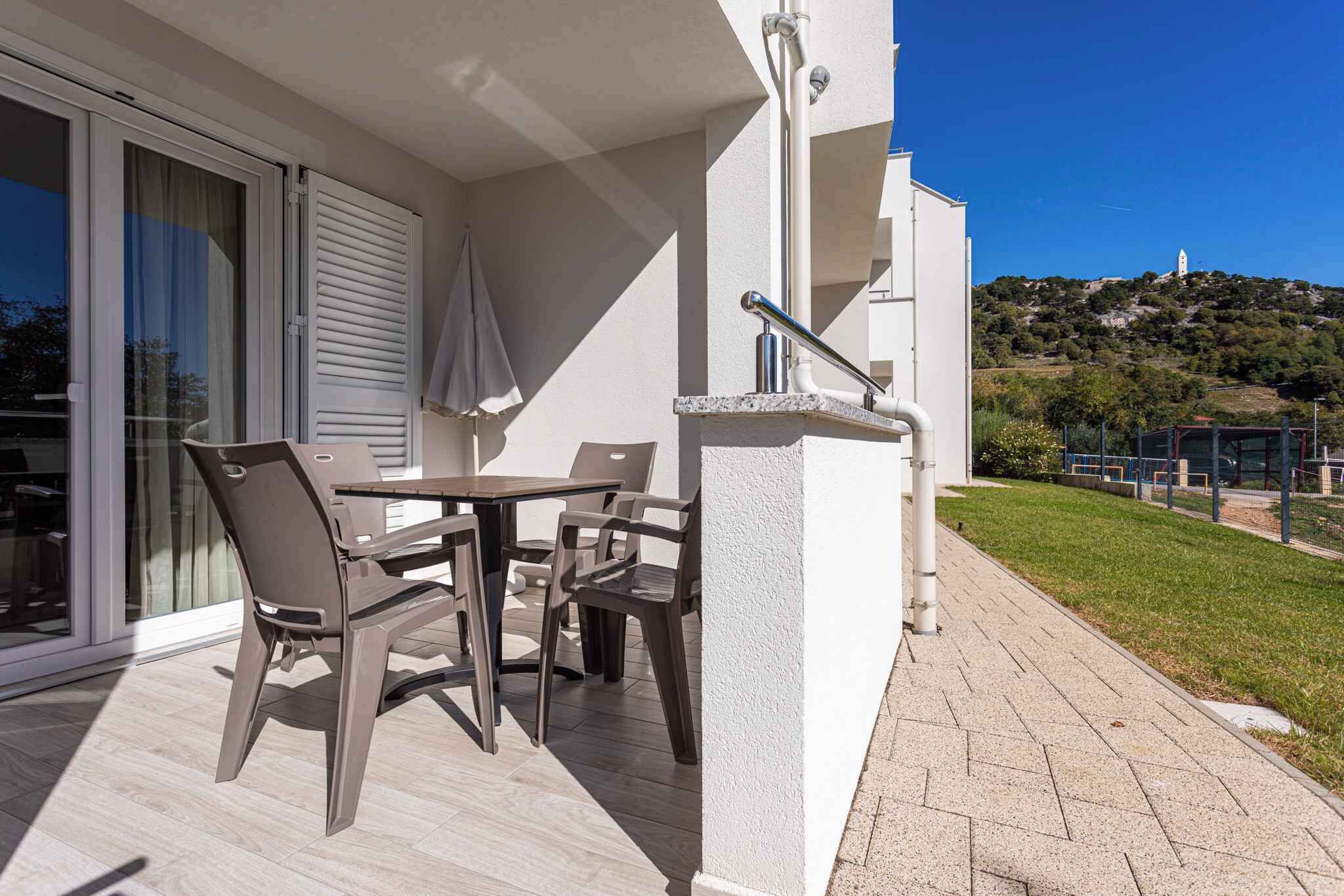 An outdoor patio with a table and chairs, a glass door, and a view of a hill with greenery and a building in the background.